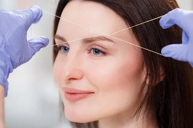 Woman receiving eyebrow threading by professional in gloves.