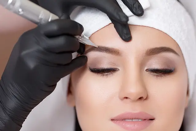 Woman receiving eyebrow microblading treatment by technician in black gloves.